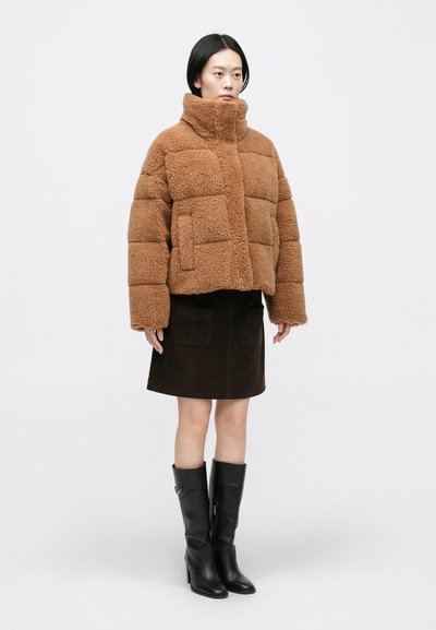 Brązowa kurtka z imitacji baranka z wysokim kołnierzem, o pudełkowym kroju i dużych przeszywanych panelach, zestawiona z ciemną zamszową spódnicą i czarnymi botkami.