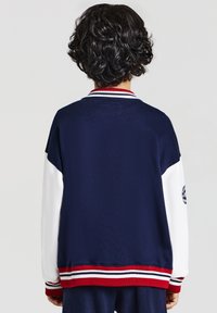 Person mit kurzen krausen Haaren, die eine marineblaue und weiße Collegejacke mit rot-weißen gestreiften Bündchen und Saum trägt, von hinten betrachtet.