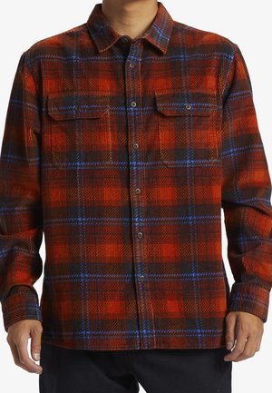 Mężczyzna w koszuli flanelowej na guziki z długim rękawem, w kratę w czerwono-czarną i niebieską kratę, z dwoma zapinanymi na guziki kieszeniami na piersi.