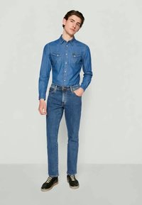 Camisa de denim en azul medio con cuello abotonado y dos bolsillos en el pecho, combinada con jeans azul medio y zapatos negros de cordones.