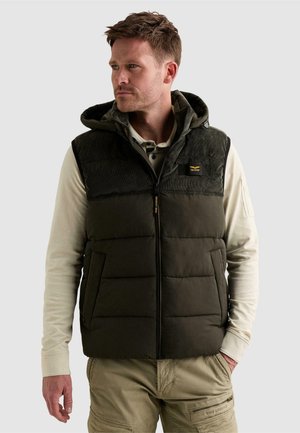 Homme portant une veste matelassée foncée avec capuche par-dessus une chemise crème à manches longues et un pantalon cargo beige, regardant vers la gauche sur un fond uni.