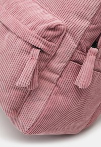 Sac à dos en velours côtelé rose avec une texture à côtes verticales, un rabat supérieur avec deux tirettes à franges et un détail de poche latérale.