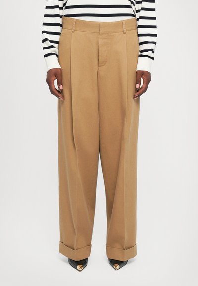 Persona vestida con pantalones de tiro alto y pierna ancha de color beige con puños, zapatos de punta negra con puntas doradas, y una camiseta de rayas en blanco y negro de mangas largas.