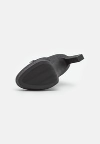 Chaussure à talon noire avec une surface texturée et une semelle en caoutchouc, présentant un bout arrondi et un talon bloc. La semelle comporte un motif ondulé pour l'adhérence.