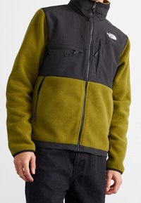 Groen fleecevest met een zwart nylon bovenstuk, voorzien van een hoge kraag, twee met ritsen afsluitbare zakken en een ritssluiting aan de voorkant.