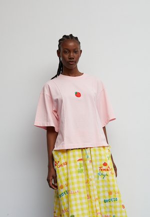 Woman with braided hair wearing a loose light pink shirt with a small tomato graphic and a yellow checkered skirt with colorful text patterns.