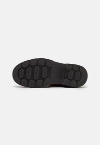 Semelle en caoutchouc noir avec un motif texturé et des rainures pour l'adhérence, comportant le nom de marque en relief "GANT" et des détails de fabrication.