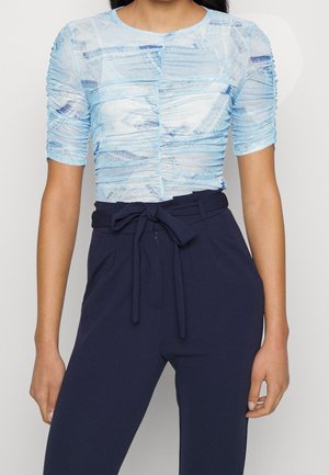 Femme portant un haut court à manches courtes froncé, bleu clair à motifs, et un pantalon taille haute bleu marine noué avec une ceinture en tissu.