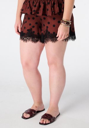 Femme portant un short marron à pois noirs avec bordure en dentelle, des sandales marron et des bracelets à un poignet, debout sur un fond blanc.