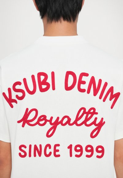 Balts kokvilnas t-krekls, kuram ir sarkans izšuvums ar tekstu: "KSUBI DENIM," "Royalty," un "SINCE 1999." Vienkāršs, ikdienišķs dizains ar apaļu kakla izgriezumu.