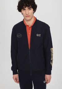 Jūras zilas krāsas džemperis ar rāvējslēdzēju, izgatavots no teksturēta auduma, zem kura redzams oranžs krekls ar apkakli. Uz krūtīm ir zelta EA7 logo un sarežģīts dizains uz piedurknes.