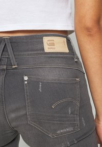Jeans de mezclilla gris con un diseño ajustado, que presentan un parche de cuero con el logotipo RAW, detalles desgastados y bolsillos traseros clásicos con costuras en contraste.