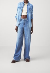 Světle modrá denimová košile na knoflíky s náprsními kapsami, v kombinaci s volnými světle modrými džínami. Převlečená přes přiléhavý krémový top.