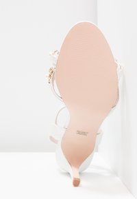 Sandale à talons blancs avec un dessus texturé, des fines lanières et ornée d'accents dorés. La semelle est lisse avec de légères rainures.