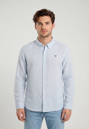 Mann trägt hellblaues langärmliges Hemd mit Knöpfen und Kragen sowie hellblaue Jeans und steht vor einem einfarbigen Hintergrund.