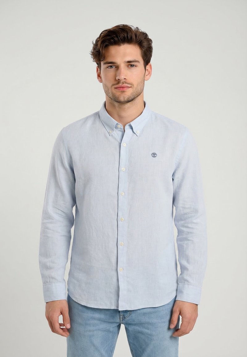 Homme portant une chemise bleu clair à manches longues avec col et boutons, ainsi qu'un jean bleu clair, debout devant un fond uni.