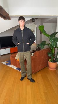 Νεαρός άνδρας με μαύρο μπουφάν και ελαιδί παντελόνι στέκεται σε σαλόνι με πορτοκαλί καναπέ, μεγάλο φυτευτό φυτό, τηλεόραση και ξύλινο πάτωμα.