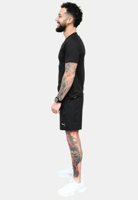Camiseta deportiva negra y pantalones cortos, con logo blanco. Zapatillas blancas. Tatuajes coloridos notables en las piernas. Peinado natural, barba.