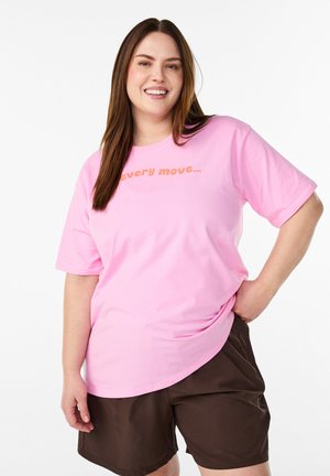 Femme souriante aux longs cheveux bruns, portant un t-shirt rose avec un texte orange et un short marron foncé, main sur la hanche, sur fond blanc.