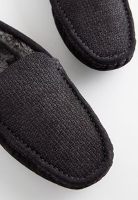 Svarta tygtofflor med vävd struktur, utrustade med ett mjukt foder och en lätt rundad tå. Försedda med sydda kanter.