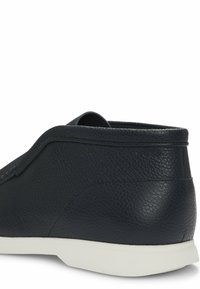 Scarpa nera in pelle senza lacci con superficie testurizzata, profilo basso e suola in gomma bianca a contrasto. Tacco arrotondato e dettagli di cucitura minimi.