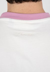 Rückenansicht von Nacken und oberem Rücken, bekleidet mit einem weißen Hemd mit pinkfarbenem Kragen und erhabener "Proclaim"-Schrift nahe am Halsausschnitt.