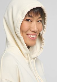 Felpa crema con cappuccio regolabile, texture morbida, vestibilità leggermente oversize, con un logo discreto sul petto e bordo del cappuccio arricciato.