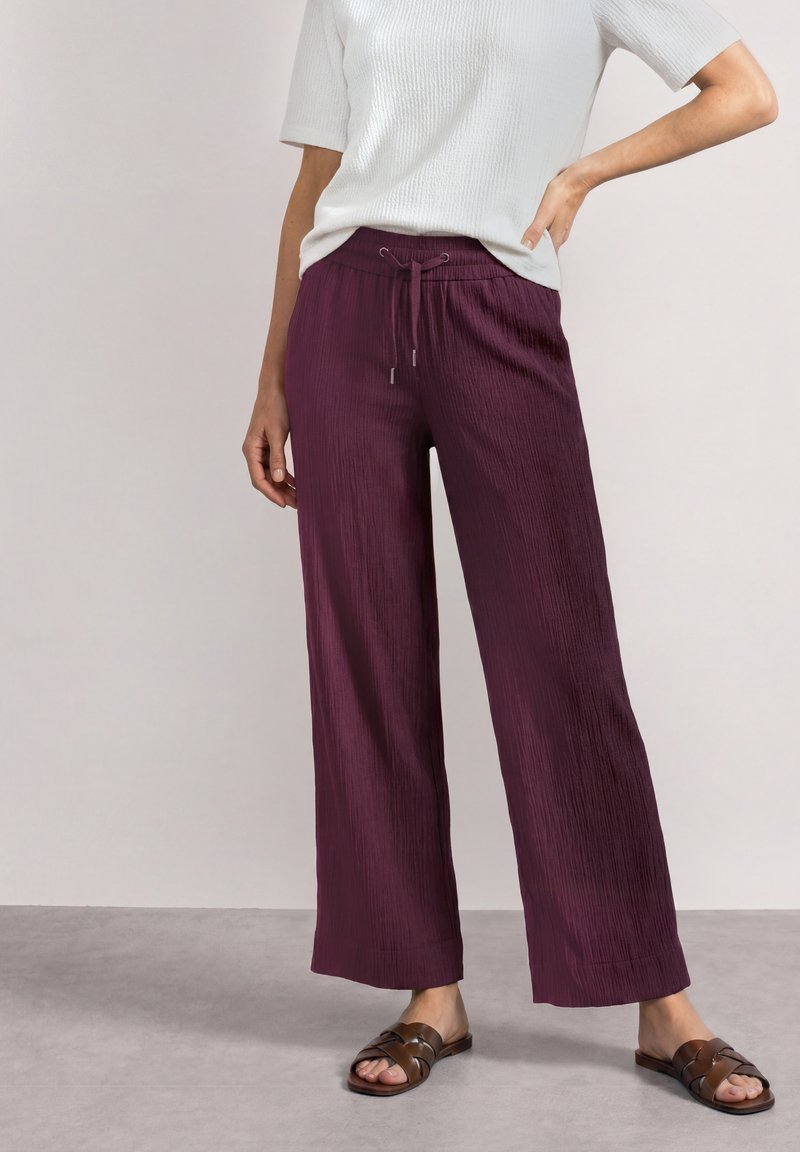 Personne portant un pantalon ample et texturé violet foncé avec une taille à cordon, un haut blanc à manches courtes et des sandales plates marron, debout sur un sol gris.