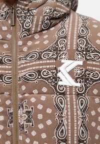 Chaqueta marrón acolchada con patrón paisley, que cuenta con un parche de logo blanco y cremallera dorada, fabricada con tela texturizada.