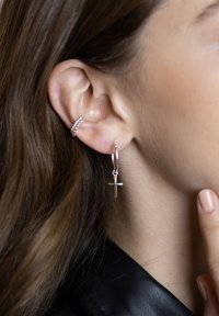 Orecchino a cerchio argento con un pendente a croce, superficie texturizzata, e un secondo orecchino a chiodo con piccoli dettagli a perline. Ingrandimento sull'orecchio.