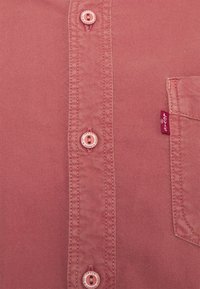 Chemise en coton de couleur rose fanée avec un col boutonné, comportant six boutons roses et une petite étiquette de marque rouge sur la poche.