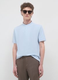 Polo a maniche corte color azzurro chiaro con colletto alla coreana, dotata di una patta con tre bottoni dello stesso tessuto. Abbinata a pantaloni sartoriali marroni.