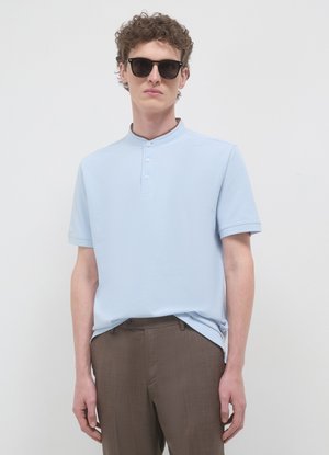 Polo a maniche corte color azzurro chiaro con colletto alla coreana, dotata di una patta con tre bottoni dello stesso tessuto. Abbinata a pantaloni sartoriali marroni.