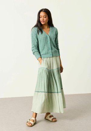 Frau steht da, trägt eine strukturierte grüne Strickjacke, einen langen plissierten Rock mit Spitzenapplikation und beige Sandalen mit Schnalle, Hände in den Taschen, schaut nach unten.