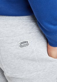 Pantalones de chándal grises con un bolsillo parche que presenta la marca "CREEKS", combinados con una camiseta de manga larga azul. Tejido suave y texturizado.