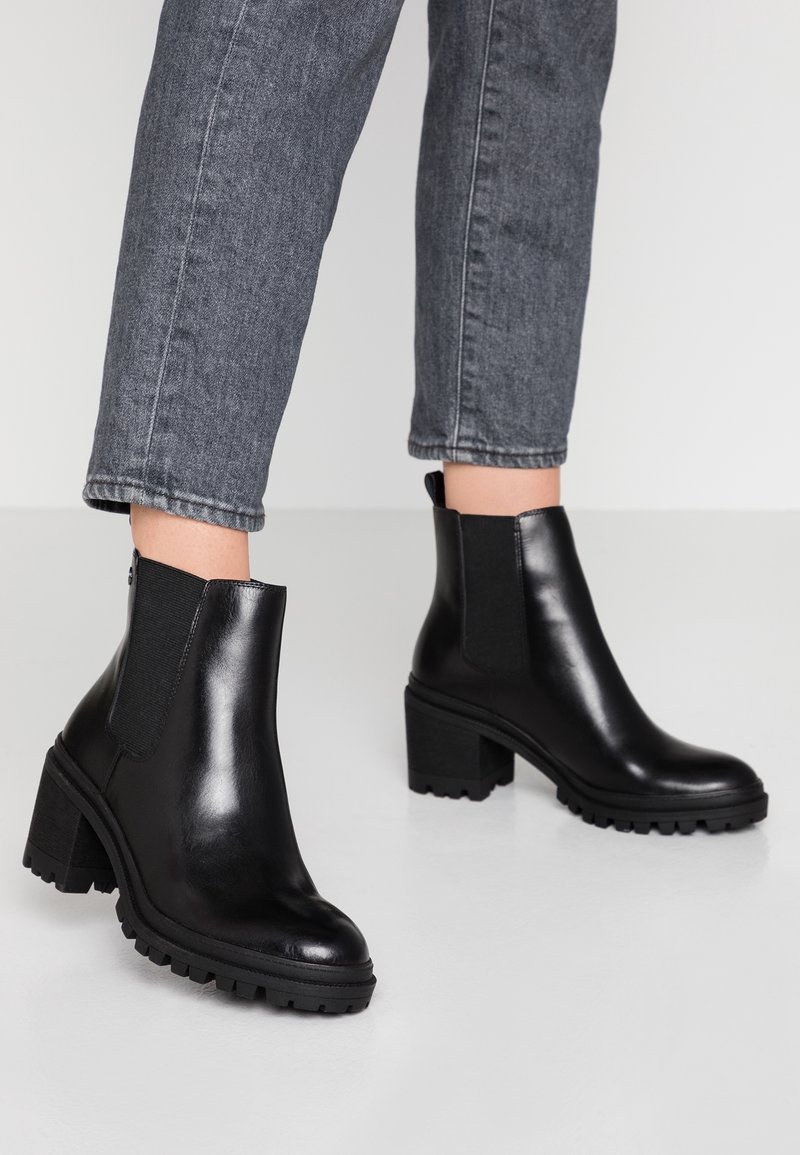 Bottines en cuir noir avec talons épais et panneaux latéraux élastiques, portées avec un jean gris foncé retroussé sur un sol clair.