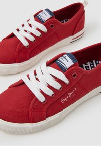 Rote Leinensneaker mit weißen Schnürsenkeln, einem Logo an der Seite und einem blau-weiß gestreiften Etikett an der Ferse. Gummisohle und strukturiertes Detail.