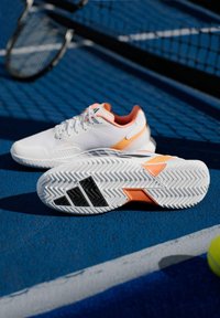 Λευκά αθλητικά παπούτσια με πορτοκαλί λεπτομέρειες, με υφασμάτινη υφή, δέσιμο με κορδόνια και μοτίβο στη λαστιχένια σόλα, κατάλληλα για τένις.