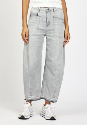 Person trägt hellgraue, weit geschnittene, kurze Jeans mit großen Fronttaschen und weißen Nike-Sneakers, steht vor einem schlichten Hintergrund.