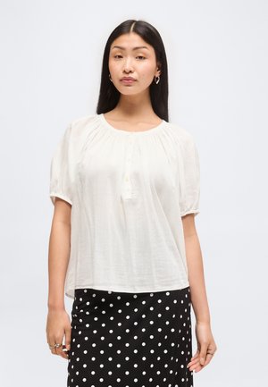 Azjatka z długimi czarnymi włosami, ubrana w białą bluzkę z krótkim rękawem i czarną spódnicę w białe kropki, stojąca na jednolitym tle.