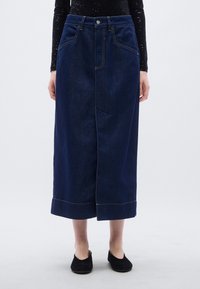 Midi φ ούστα από τζιν, σκούρο μπλε, ψηλόμεση, με πέντε τσέπες, αντίθετη ραφή, πλαϊνή σχισμή, τελείωμα με ρέλι, φορεμένη με μαύρα φλατ παπούτσια.