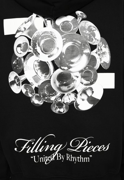 Skupina reflektivnih zvona od mesinga s natpisom "Filling Pieces 'United By Rhythm'" na crnoj tkanini.