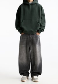 Felpa verde con cappuccio e tasca frontale, abbinata a jeans in denim grigi con gamba larga che sfumano da scuri a chiari. Sneakers grigie.