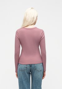 Γυναίκα με μακριά ξανθά μαλλιά, που φοράει εφαρμοστή λεβαντίνι μακρυμάνικη μπλούζα και μπλε τζιν, με την πλάτη στραμμένη προς λευκό φόντο.