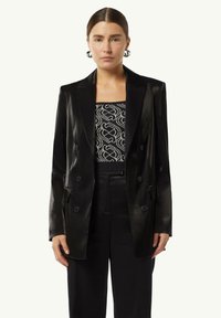 Black satin double-breasted blazer with peak lapels, single front pocket, and two buttons. Worn over a patterned black and white top.