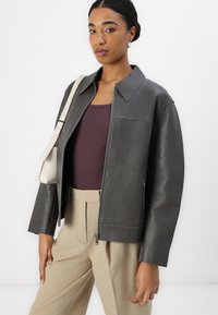 Graue Lederjacke mit großem Kragen, Reißverschluss und zwei vorderen Taschen. Getragen über einem gerippten braunen Oberteil und kombiniert mit beigen Hosen.