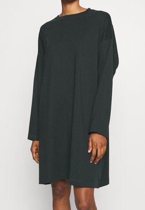 Mannequin portant une robe noire à manches longues, ample et longueur genou, sur fond blanc uni.