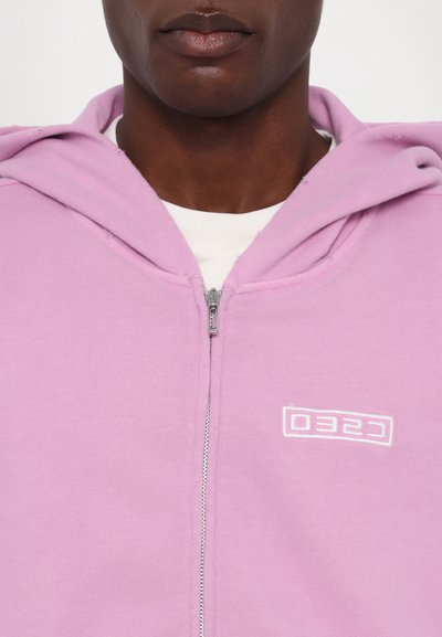 Light pink zip-up hoodie with a white embroidered logo on the left chest. Features a front zipper and a soft, textured fabric.