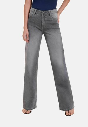 Persoon draagt grijze high-waisted jeans met wijde pijpen, gecombineerd met een navy top en zwarte open-toe sandalen tegen een effen achtergrond.