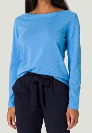 Femme portant un haut à manches longues bleu clair texturé, rentré dans un pantalon noir taille haute avec une ceinture à nouer à l'avant.